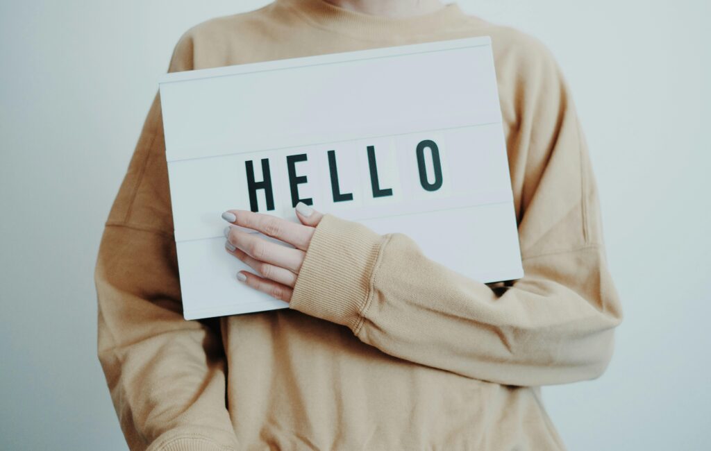 pexels photo 4439411 4439411 A person in beige sweater holding a 'HELLO' sign against a light background.