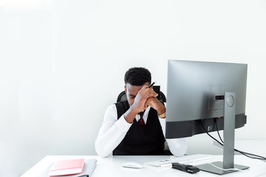 A businessman sitting at a desk, appearing stressed while working at a computer in a bright office.